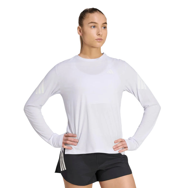 En kvinde iført en Adidas Adi365 Running Iconic langærmet trøje til kvinder i farven Fropur/hvid og sorte shorts med hvide striber står med hænderne på hofterne og kigger til siden. Tøjet er udstyret med Climacool-teknologi, der sikrer åndbar komfort.
