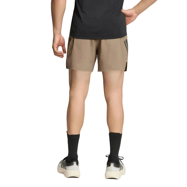 Der ses en person set bagfra, iført en sort T-shirt, Adidas Adi365 Running Iconic-shorts til mænd i farven Khasix/Black, sorte sokker og hvide sportssko. Den ene hånd hviler på hoften foran en ensfarvet hvid baggrund.