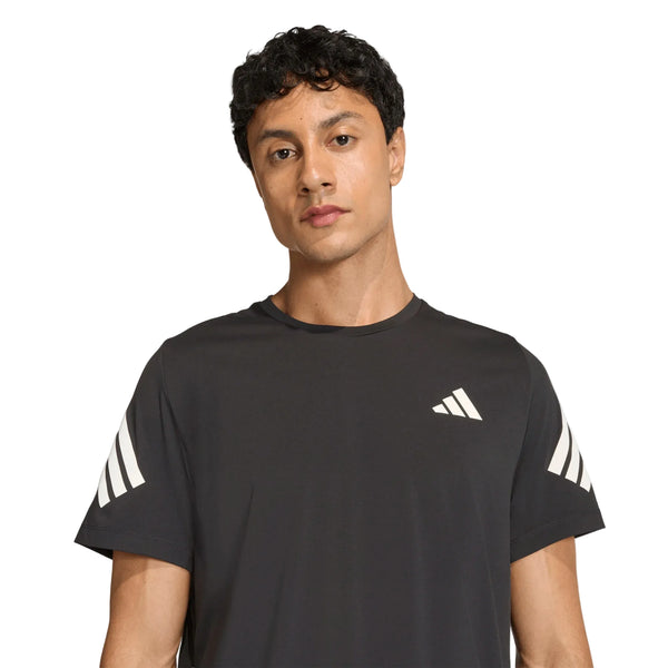 En mand med kort, krøllet hår har en Adidas Adi365 Running Iconic T-shirt, Herre, Sort på – en sort løbetrøje til mænd med hvide striber på ærmerne og et lille hvidt geometrisk logo på brystet, udviklet med adidas Climacool-teknologi – og står foran en ensfarvet hvid baggrund.