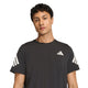 En mand med kort, krøllet hår har en Adidas Adi365 Running Iconic T-shirt, Herre, Sort på – en sort løbetrøje til mænd med hvide striber på ærmerne og et lille hvidt geometrisk logo på brystet, udviklet med adidas Climacool-teknologi – og står foran en ensfarvet hvid baggrund.