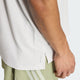 En person står med armen afslappet ned langs siden og har en Adidas Adi365 Running Iconic T-shirt til mænd i farven Greone/White på samt lysegrønne shorts med hvide striber. Fokus er på den nederste del af Adidas-løbet-T-shirten og personens hånd.