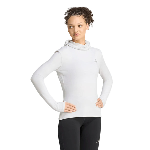 En person poserer iført Adidas Adi365 Running Winterized Long Sleeve træningsoverdel til kvinder i farven Greone, der har et diskret logo på brystet og er kombineret med sorte leggings. Med hænderne på hofterne og hovedet let vendt til siden fremviser vedkommende denne stilfulde langærmede træningsoverdel – perfekt til træning i køligt vejr.
