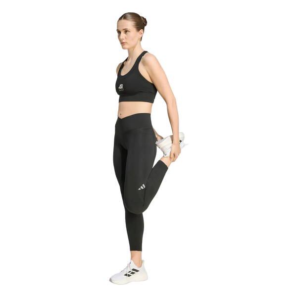 En kvinde iført sorte Adidas Adi365 Seasonal 7/8-tights står på det ene ben og holder den anden fod bag sig for at strække låret. Hun har hvide sneakers på og ser fremad med et neutralt udtryk foran en ensfarvet hvid baggrund.