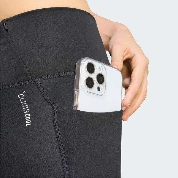 En person lægger en smartphone med et gennemsigtigt cover ned i sidelommen på et par Adidas Adi365 Seasonal 7/8-tights, dame, sort. Kun den nedre del af kroppen og hånden er synlige på en ensfarvet lys baggrund.