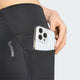 En person lægger en smartphone med et gennemsigtigt cover ned i sidelommen på et par Adidas Adi365 Seasonal 7/8-tights, dame, sort. Kun den nedre del af kroppen og hånden er synlige på en ensfarvet lys baggrund.