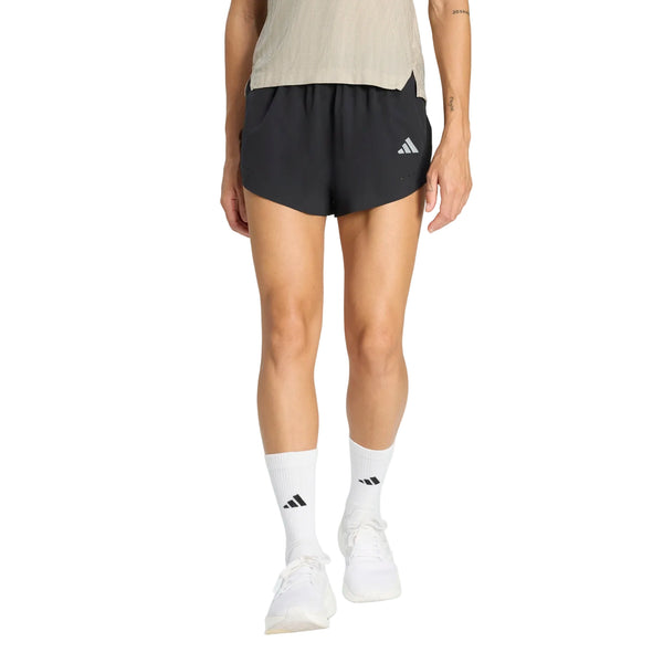 En person står med ansigtet vendt fremad, så man kun kan se den nederste del af kroppen. Personen har en lys beige skjorte på, sorte Adidas Adi365 Climacool+2 Pocket Shorts 5" (Dame), hvide Adidas-sokker og hvide sportssko.