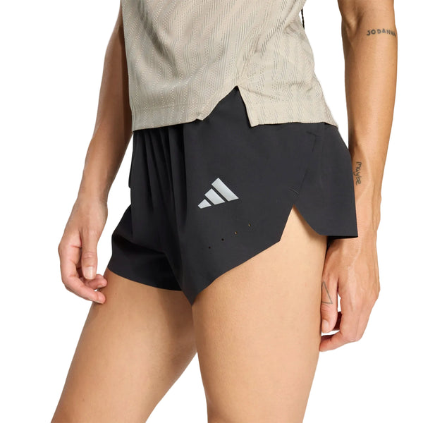 En person iført en beige kortærmet skjorte og et par sorte Adidas Adi365 Climacool+2 Pocket Shorts 5" til kvinder med et reflekterende logo står med den ene hånd afslappet ned langs siden.