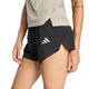 En person iført en beige kortærmet skjorte og et par sorte Adidas Adi365 Climacool+2 Pocket Shorts 5" til kvinder med et reflekterende logo står med den ene hånd afslappet ned langs siden.