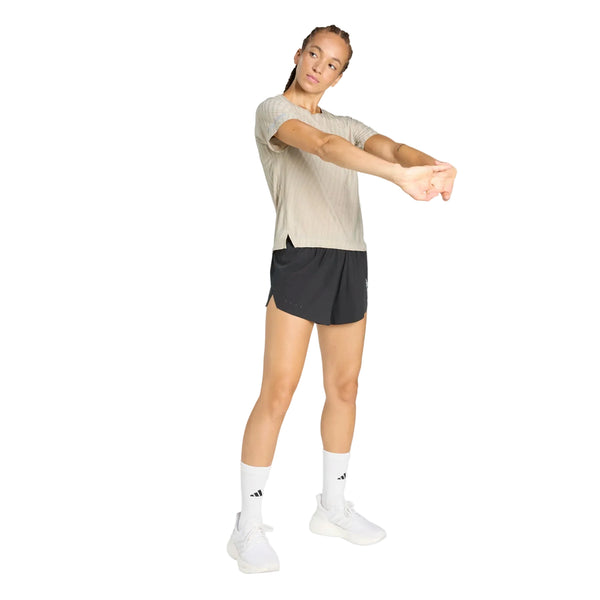 En person i sportstøj strækker sig. Personen har en lys beige T-shirt på, sorte Adidas Adi365 Climacool+2 Pocket Shorts 5" til kvinder, hvide sokker med sorte Adidas-logoer og hvide sportssko på, alt sammen på en hvid baggrund.