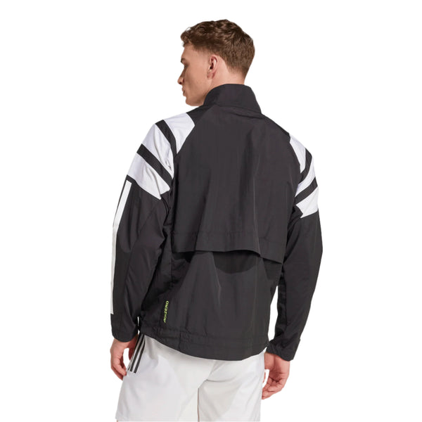En mand iført Adidas Adizero A Jkt M løbejakke i sort med geometriske mønstre står med ansigtet vendt mod kameraet sammen med hvide shorts. Den stilfulde jakke står i kontrast til den ensfarvede hvide baggrund.