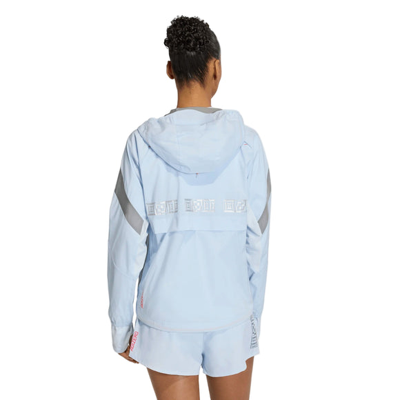 En kvinde med kort, krøllet hår står med ryggen til kameraet iført en lyseblå Adidas Adizero Hakone Ekiden Jacket, Dame (Crsk/Silvmt), som har refleksbånd på ærmerne og et hvidt bånd hen over ryggen.