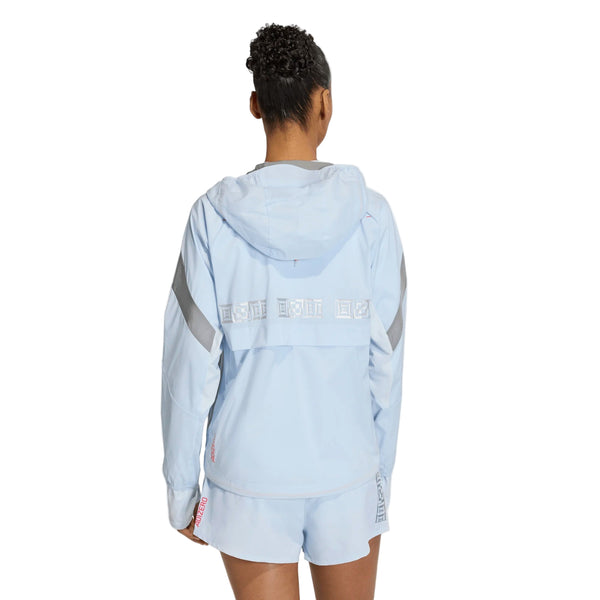 En kvinde med kort, krøllet hår står med ryggen til kameraet iført en lyseblå Adidas Adizero Hakone Ekiden Jacket, Dame (Crsk/Silvmt), som har refleksbånd på ærmerne og et hvidt bånd hen over ryggen.
