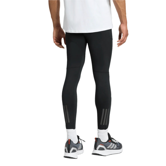 En person står med ryggen til og er iført en hvid skjorte og Adidas Otr Bin Tig løbetights, Herre, Black med reflekterende lægstriber, hvide sokker og grå sneakers med hvide såler. Kun den nederste halvdel af kroppen er synlig.