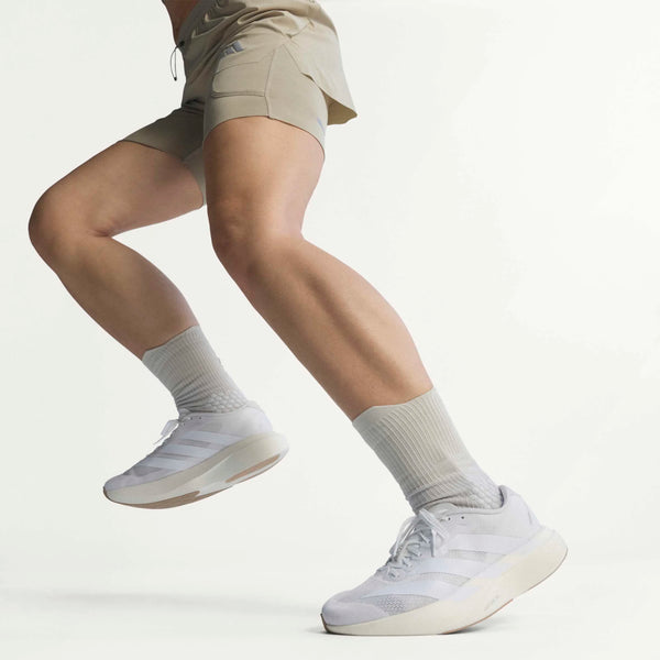 En kvinde midt i et spring, vist fra benene og ned, er iført beige sportsshorts og Adidas Adizero Evo SL Dame Ftwwht/Ftwwht/Warsan-sneakers med lysegrå strømper på en almindelig hvid baggrund.