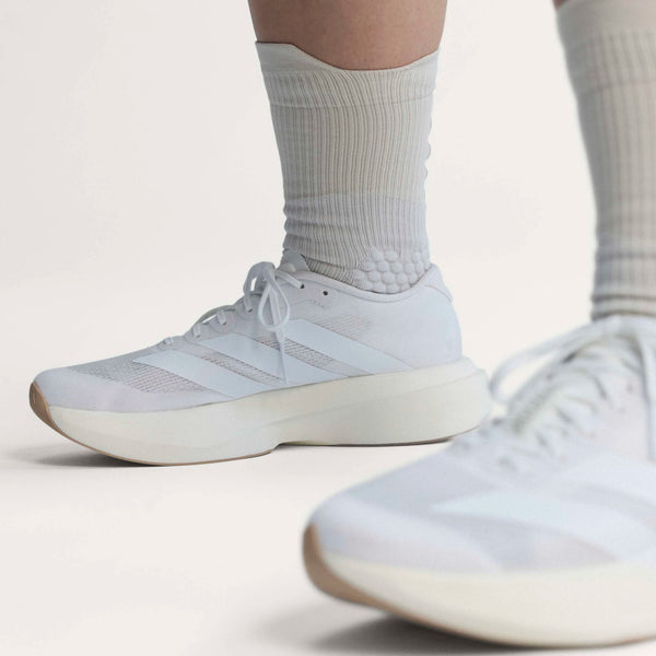 En person iført hvide, ribbede sokker og Adidas Adizero Evo SL Dame-sko i Ftwwht/Ftwwht/Warsan med en tyk LIGHTSTRIKE PRO-sål og tre striber står på en lys flade med fokus på skoene og underbenene.
