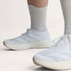 En person iført hvide, ribbede sokker og Adidas Adizero Evo SL Dame-sko i Ftwwht/Ftwwht/Warsan med en tyk LIGHTSTRIKE PRO-sål og tre striber står på en lys flade med fokus på skoene og underbenene.