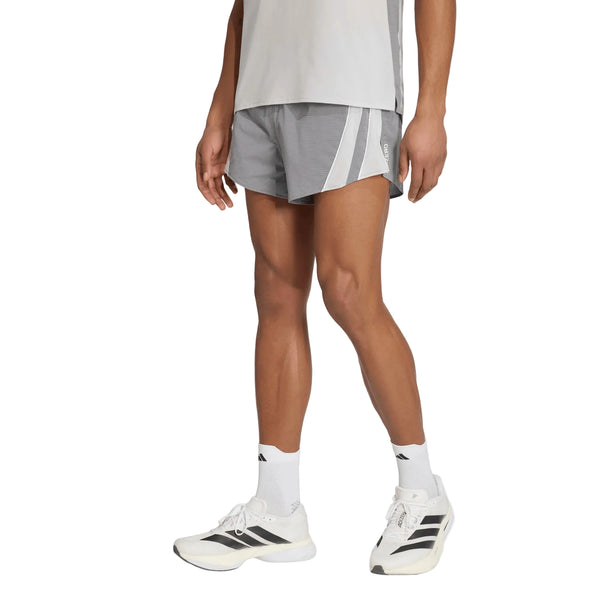 En person står foran en ensfarvet hvid baggrund iført en lysegrå skjorte, Adidas Adizero Archive 5″ løbeshorts til mænd i farven Grethr/Greone – en moderne model af herre-løbeshorts – kombineret med hvide strømper og hvide sportssko med sorte striber, hvilket perfekt fremhæver moderne herre-løbetøj.