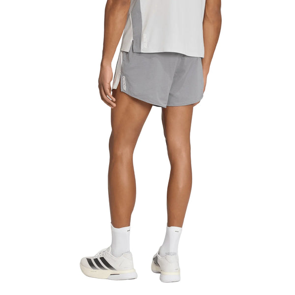 En person står med ryggen til kameraet og er iført en lysegrå skjorte, Adidas Adizero Archive 5″ løbeshorts til mænd i farven Grethr/Greone, hvide strømper og hvide sportssko med sorte striber. Baggrunden er helt hvid.