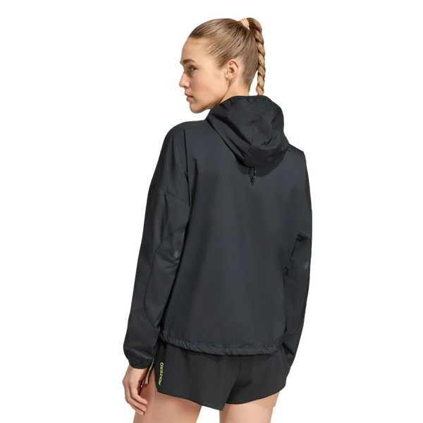 En kvinde med lysebrunt, flettet hår er iført en sort Adidas Adizero Running Blaze-jakke og sorte shorts. Hun står med ryggen til kameraet og hovedet vendt til siden foran en ensfarvet hvid baggrund.