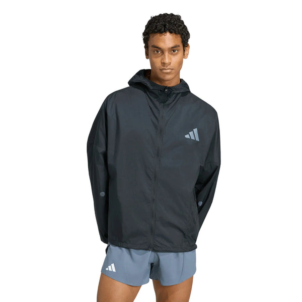 En person står med ansigtet vendt fremad iført en Adidas Adizero Running Blaze-jakke, Herre, i sort, kombineret med lyseblå sportsshorts. Både jakken og shortsene har et lille hvidt Adidas-logo. Baggrunden er ensfarvet hvid.