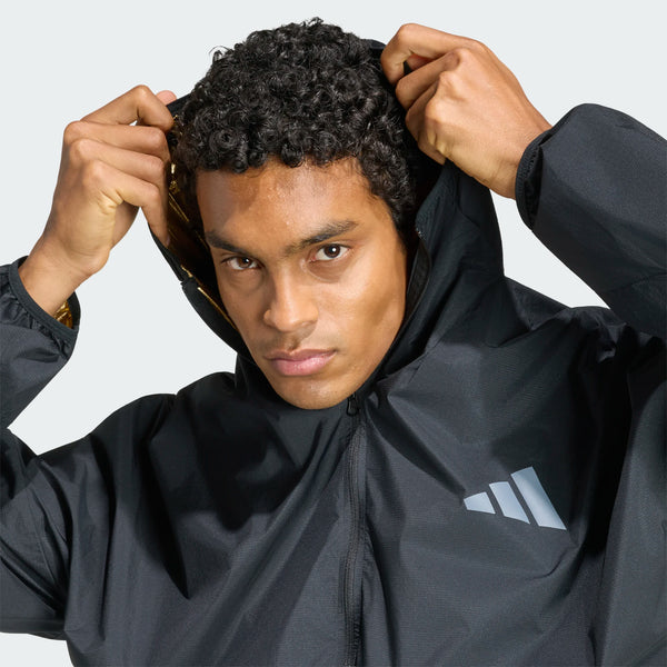 En person med krøllet hår har en Adidas Adizero Running Blaze-jakke (herre, sort) på og holder hætten oppe med begge hænder. Denne løbejakke til mænd har et hvidt, geometrisk Adidas-logo på venstre bryst. Baggrunden er ensfarvet og lys.