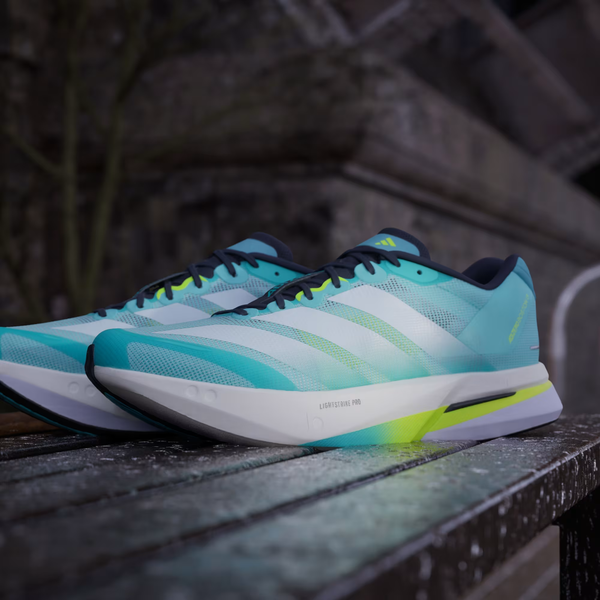 Et par Adidas Adizero Boston 13 herre løbesko i Flash Aqua/Cloud White/Lucid Lemon hviler på en træbænk udendørs med en sløret betonkonstruktion i baggrunden.