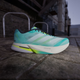 Adidas Adizero Boston 13 herre løbesko i Flash Aqua/Cloud White/Lucid Lemon vises på en udendørs belagt overflade med slørede murstensbygninger og metalgelændere i baggrunden.