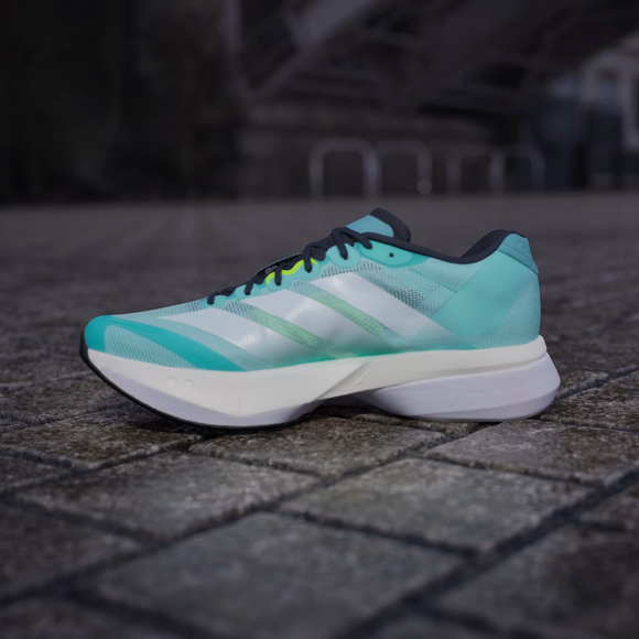 En Adidas Adizero Boston 13 herre løbesko i Flash Aqua/Cloud White/Lucid Lemon har en tyk hvid sål og hviler på en stenbelagt overflade udendørs med en sløret bybaggrund.
