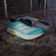 Et par Adidas Adizero Boston 13 herre løbesko i Flash Aqua/Cloud White/Lucid Lemon er placeret på en våd, stenbelagt jord ved siden af en murstensvæg og metalcykelstativer.