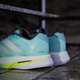 Et nærbillede af Adidas Adizero Boston 13 herre løbesko i Flash Aqua/Cloud White/Lucid Lemon, der har et stilfuldt blågrønt og hvidt design med gule detaljer, placeret på en stenoverflade ved siden af et metalgelænder.