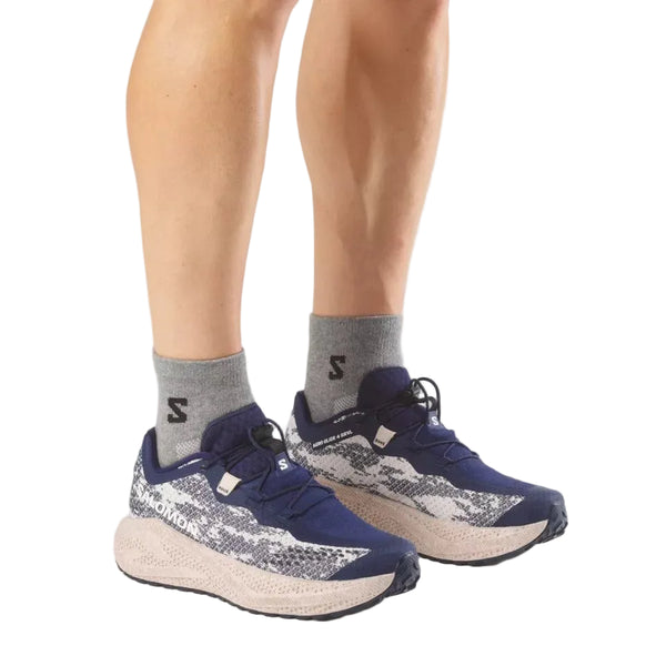 En person iført grå sokker og Salomon Aero Glide 4 Grvl herresko i farvekombinationen Mar Bl/Slv Cl/Wh står på en hvid baggrund. Man kan kun se underbenene og fødderne, klar til at dæmpe stød på grusveje.