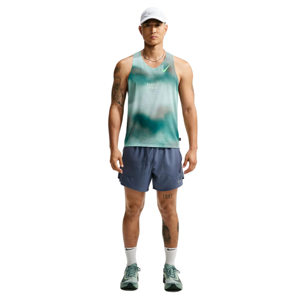En mand står med ansigtet vendt fremad, klædt i Nike-løbetøj til mænd: en hvid kasket, Nike AeroSwift Running Singlet til mænd i farven Bright Spruce/Green tie-dye, blå sportsshorts med Dri-FIT ADV-teknologi, hvide sokker og grønne løbesko. Han har tatoveringer på venstre arm og højre ben. Baggrunden er ensfarvet hvid.