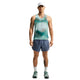 En mand står med ansigtet vendt fremad, klædt i Nike-løbetøj til mænd: en hvid kasket, Nike AeroSwift Running Singlet til mænd i farven Bright Spruce/Green tie-dye, blå sportsshorts med Dri-FIT ADV-teknologi, hvide sokker og grønne løbesko. Han har tatoveringer på venstre arm og højre ben. Baggrunden er ensfarvet hvid.