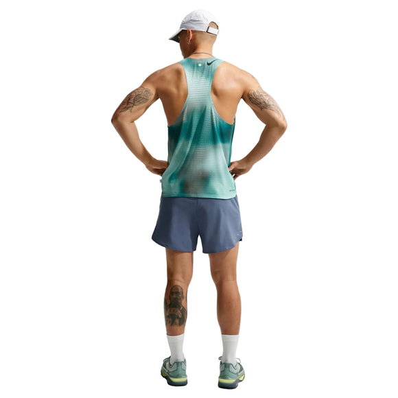 En mand står med ryggen til, hænderne i siden, iført en hvid kasket og Nike AeroSwift Running Singlet, Herre, i farven Bright Spruce/Green – et eksklusivt stykke løbetøj til mænd – kombineret med blå shorts, hvide sokker og løbesko. Han har tatoveringer på armene og venstre ben. Baggrunden er hvid.