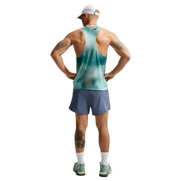 En mand står med ryggen til, hænderne i siden, iført en hvid kasket og Nike AeroSwift Running Singlet, Herre, i farven Bright Spruce/Green – et eksklusivt stykke løbetøj til mænd – kombineret med blå shorts, hvide sokker og løbesko. Han har tatoveringer på armene og venstre ben. Baggrunden er hvid.