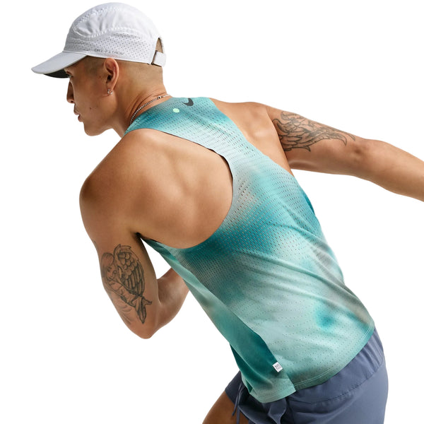En person iført en hvid kasket, blå shorts og en Nike AeroSwift løbetrøje til herre i farven Bright Spruce/Green med Dri-FIT ADV-teknologi ser ud til at løbe eller træne. Billedet er taget bagfra og viser tatoveringer på begge arme – hvilket fremhæver det stilfulde og funktionelle løbetøj fra Nike til mænd.