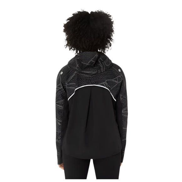 En kvinde med krøllet hår, set bagfra, er iført Asics Limited Series Lite-Show Jacket i Performance Black. Den har et hvidt geometrisk mønster, en reflekterende vandret stribe bagpå, lange ærmer og en afslappet pasform.