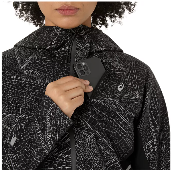 Iført en Asics Limited Series Lite-Show Jacket, Dame, i Performance Black med et geometrisk prikket design og lynlås foran, placerer en person en sort smartphone i den indvendige brystlomme. Vedkommendes ansigt er delvist synligt.