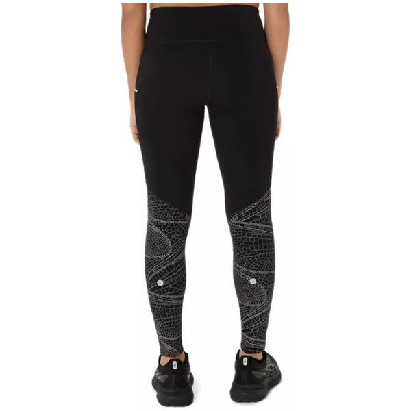 En kvinde ses bagfra iført Asics Limited Series Lite-Show Tight, Dame i Performance Black med hvide geometriske mønstre, parret med sorte sneakers og en sort top, stående mod en almindelig hvid baggrund.
