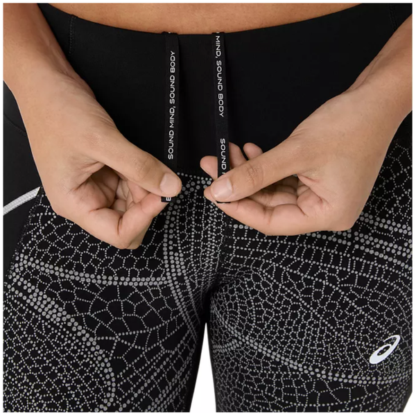 En person justerer snoren på Asics Limited Series Lite-Show Tight, Dame, Performance Black leggings med et hvidt, prikket geometrisk mønster og ASICS-logoet nær hoften. Snorene viser SOUND MIND SOUND BODY.