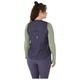 En person med krøllet hår står med ryggen til kameraet mod en hvid baggrund iført en Asics Nagino Run Padded Gilet til kvinder i Indigo Fog over en grøn langærmet top og matchende leggings.