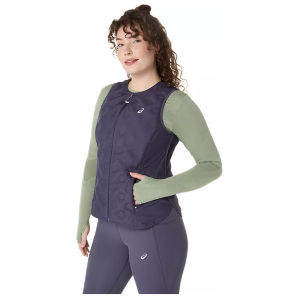 En kvinde med krøllet brunt hår bærer Asics Nagino Run Padded Gilet i Indigo Fog over en lysegrøn langærmet top og matchende marineblå leggings, mens hun står på en almindelig hvid baggrund.