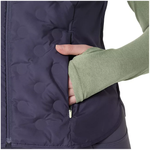 Iført en lysegrøn langærmet skjorte og en mørklilla Asics Nagino Run Padded Gilet til kvinder i Indigo Fog står en person med højre hånd i vestelommen. Gilet'en har lynlås foran og cirkulære quiltningsmønstre.