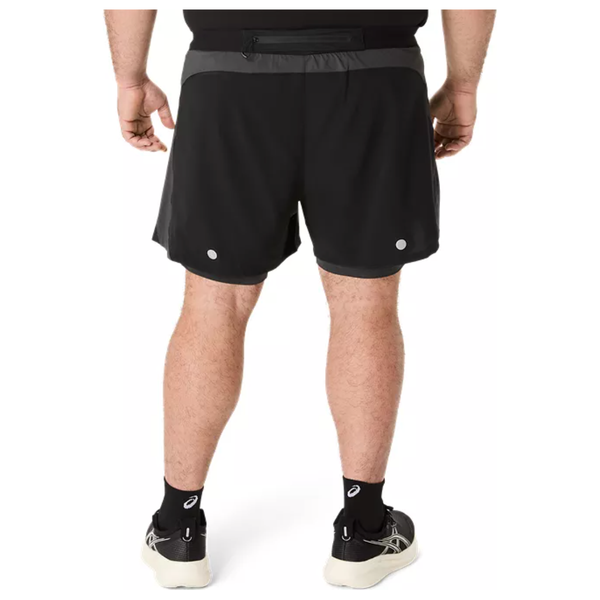 En person står med ryggen til kameraet iført Asics Road 2-N-1 5In Shorts i Performance Black/Graphite Grey, sorte ankelsokker og hvide sportssko på en almindelig hvid baggrund.