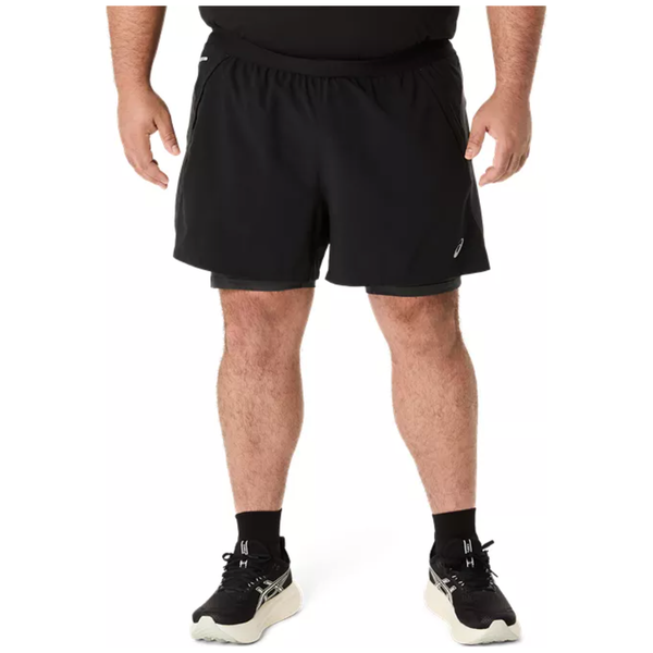 En person iført Asics Road 2-N-1 5In Shorts i Performance Black/Graphite Grey, sorte sokker og hvide løbesko står med ansigtet fremad og hænderne i siden. Kun underkroppen er synlig mod en hvid baggrund.