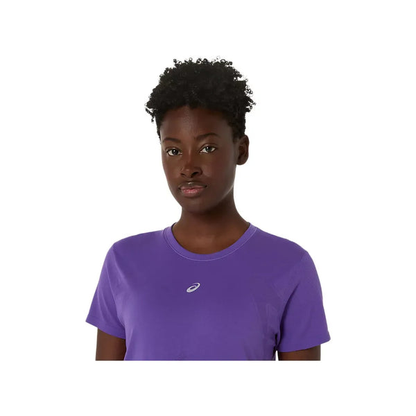 En kvinde med kort krøllet hår er iført Asics Road Seamless SS Top i Edo Purple, åndbart træningstøj, og kigger ind i kameraet mod en almindelig hvid baggrund.
