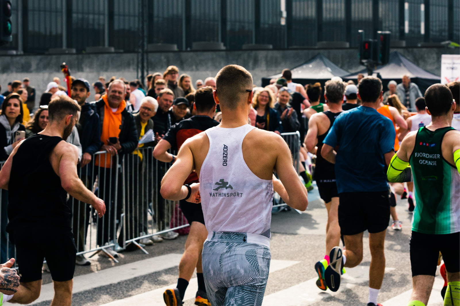 Løbere, der deltager i et landevejsløb, passerer forbi en flok jublende tilskuere bag metalbarrierer på en gade i byen. Fokus er på en løber iført en lys tanktop med kort hår.
