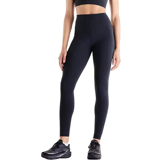 En person iført en sort sports-BH, et par New Balance Athletics Heat High Rise Legging 27″ til kvinder i sort samt sorte sportssko står foran en hvid baggrund. Armene hænger afslappet ned langs siden, og ansigtet kan ikke ses.