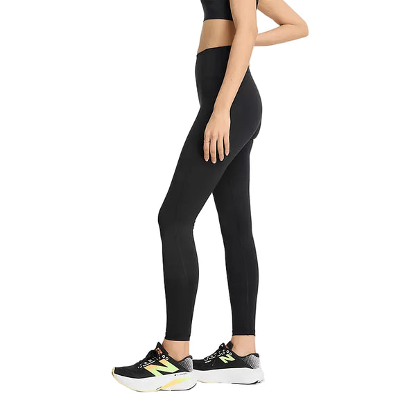 En kvinde står set i profil iført en sort sports-BH, sorte New Balance Athletics Heat High Rise Legging 27″ (dame) og sorte sportssko med gule og hvide detaljer. Leggingsene er udstyret med NB HEAT-teknologi, der sikrer øget komfort under træningen. Baggrunden er ensfarvet hvid.