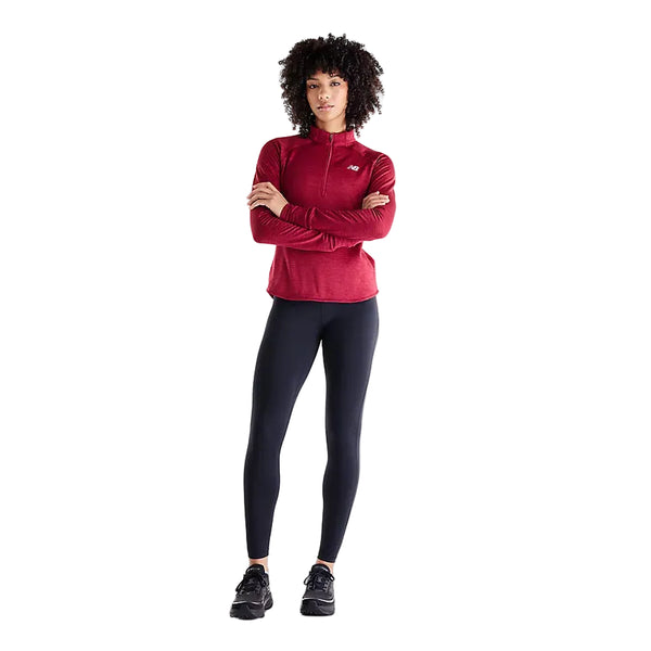 En kvinde med kort, krøllet hår står med ansigtet vendt fremad og armene over kors. Hun er iført en rød langærmet sportstrøje, sorte New Balance Athletics Heat High Rise Legging 27″ med NB HEAT-teknologi samt sorte sneakers, alt sammen på en ensfarvet hvid baggrund.
